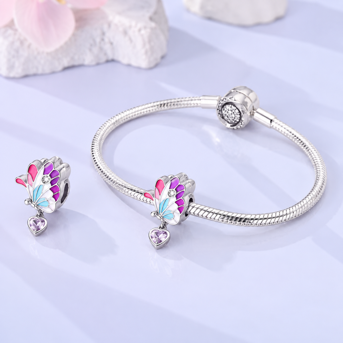 A silver-toned snake-bangle bracelet with a butterfly charm and matching butterfly stud earring, displayed on a light lavender surface with soft decorative stones in the background.